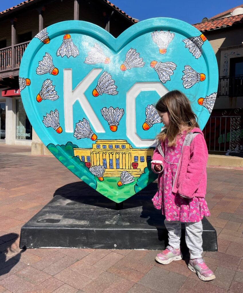 A girl looking at a giant heart with KC on it, decorated by an artist with a painting of the Nelson-Atkins Art Museum