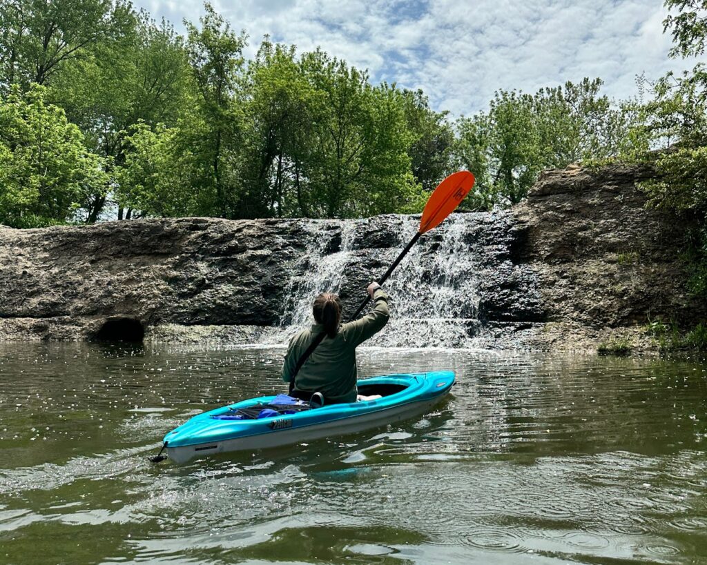 a woman paddling in a kayak in front of Cedar Creek Waterfall at Lake Olathe