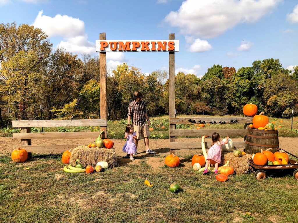 girls looking at pumpkins with their dad in front of a sign that says pumpkins