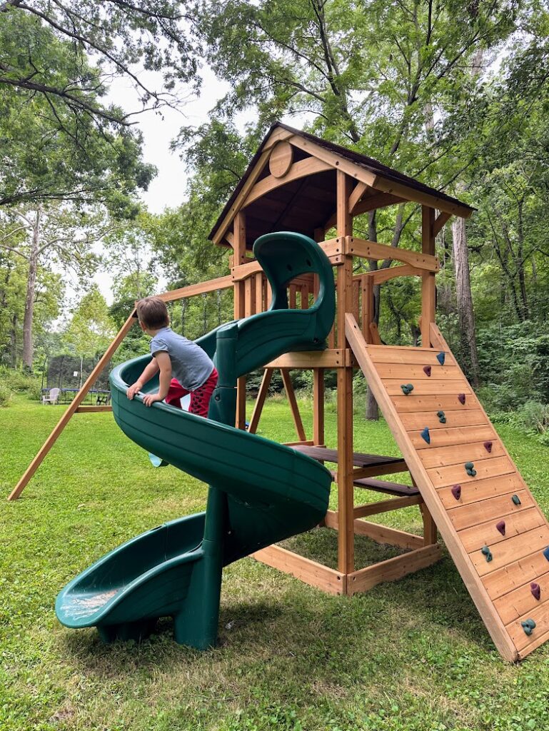 A boy playing on the slide of a swing set with a large rock climbing wall