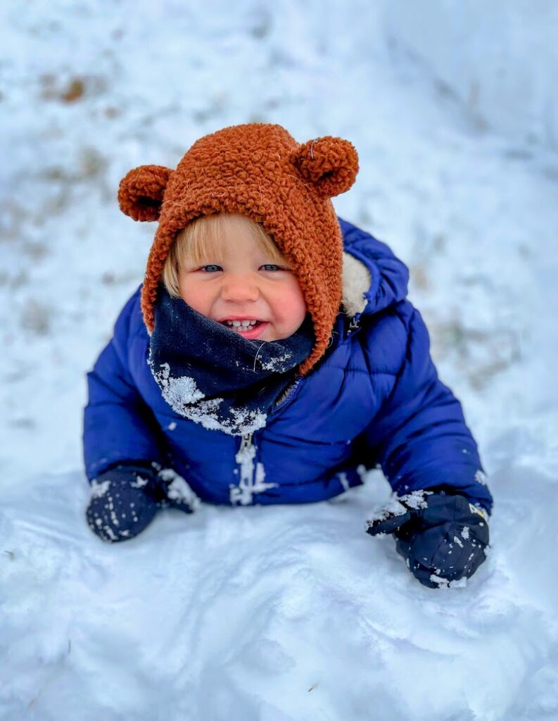 A little boy smiling in the snow with his hood up and bear ears on it 