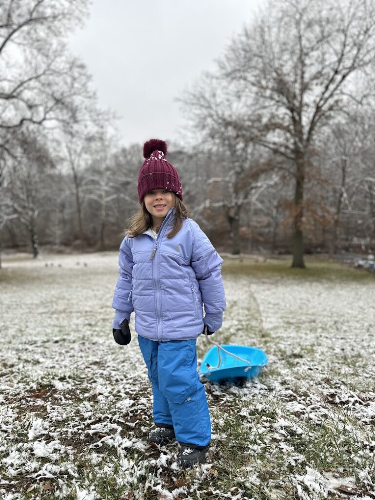 A girl standing in light snow wearing ThermKids and pulling a sled 