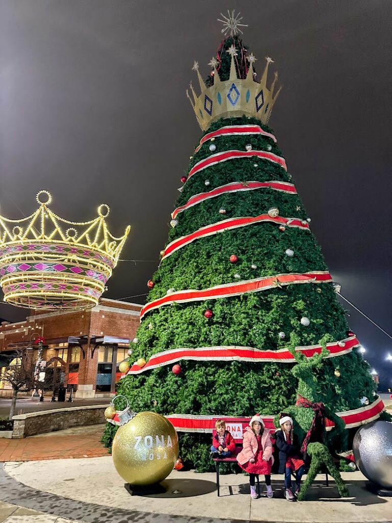 Three kids sitting on a bench in front of a huge Christmas tree at Zona Rosa in Kansas City 